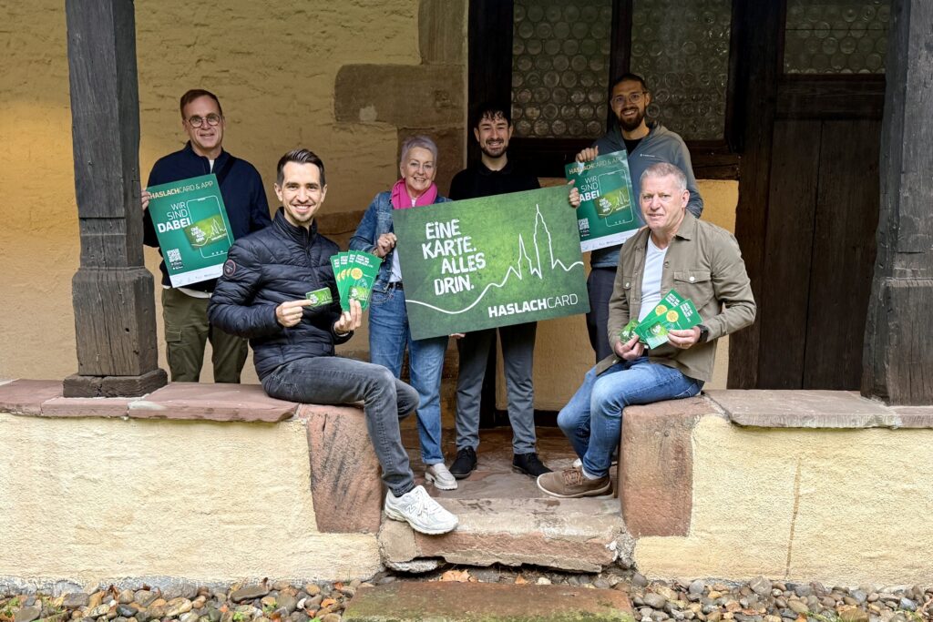 Sechs Personen stehen und sitzen auf einer alten Steintreppe vor einem historischen Gebäude. Sie halten grüne Plakate und Flyer mit der Aufschrift „HaslachCard – Eine Karte. Alles drin.“ und „Wir sind dabei“ in den Händen. Alle lächeln in die Kamera und präsentieren die neue HaslachCARD.